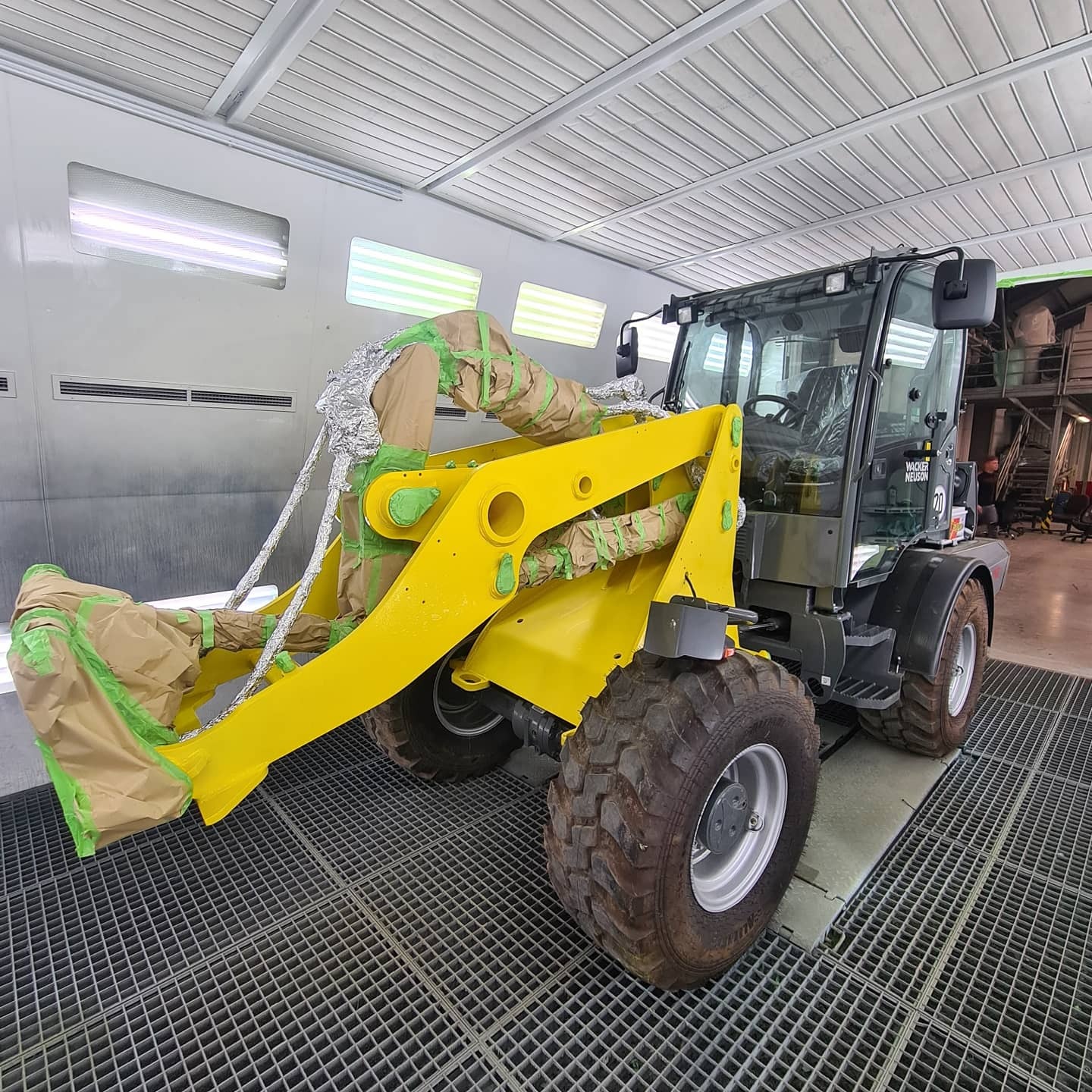 Gelber Radlader (Bagger) in einer Lackierkabine, vorbereitet für die Neulackierung mit braunem Papier und grünem Klebeband abgedeckt.