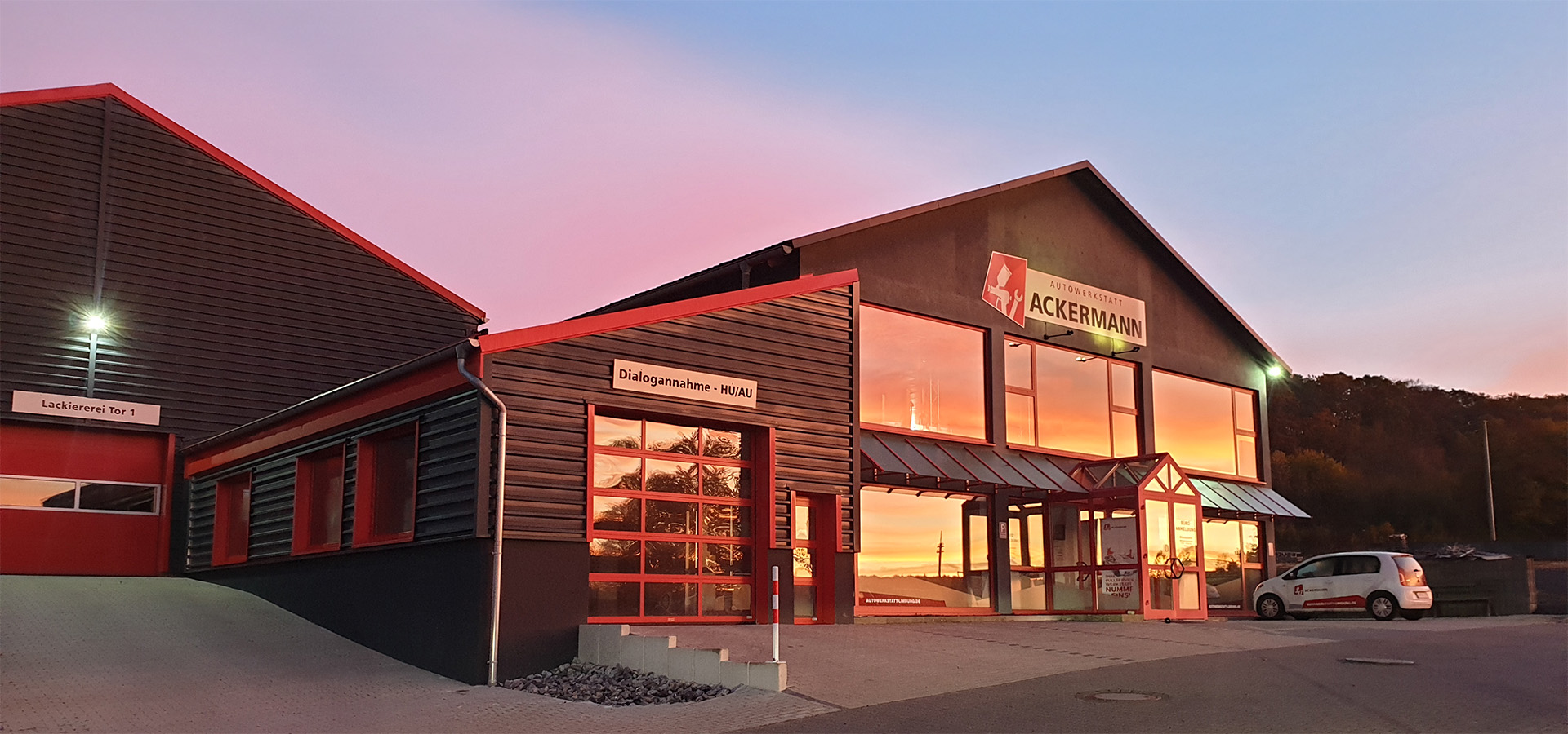 Autowerkstatt Ackermann in Merenberg-Allendorf: Gebäude bei Sonnenuntergang mit Firmenschild, Eingangsbereich und angegliedertem Lackiererei-Gebäude.